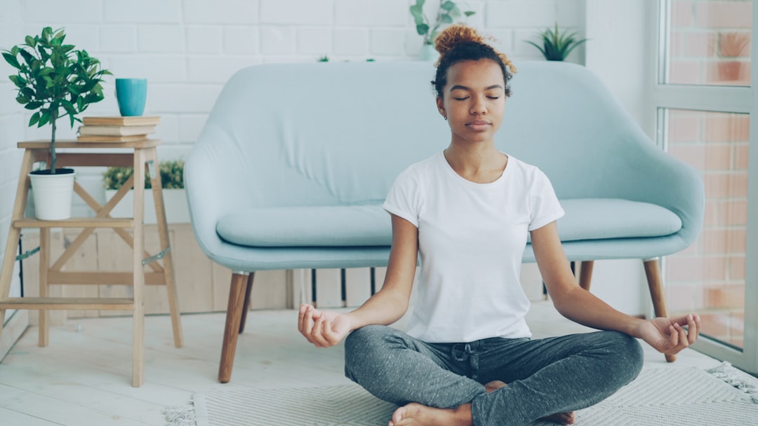 Pretty African American girl is sitting on floor in lotus position with hands on knees and meditating, enjoying relaxation and tranquility. Yoga, young people and home concept.