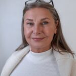 Senior woman with glasses smiling warmly in a cozy white sweater indoors.