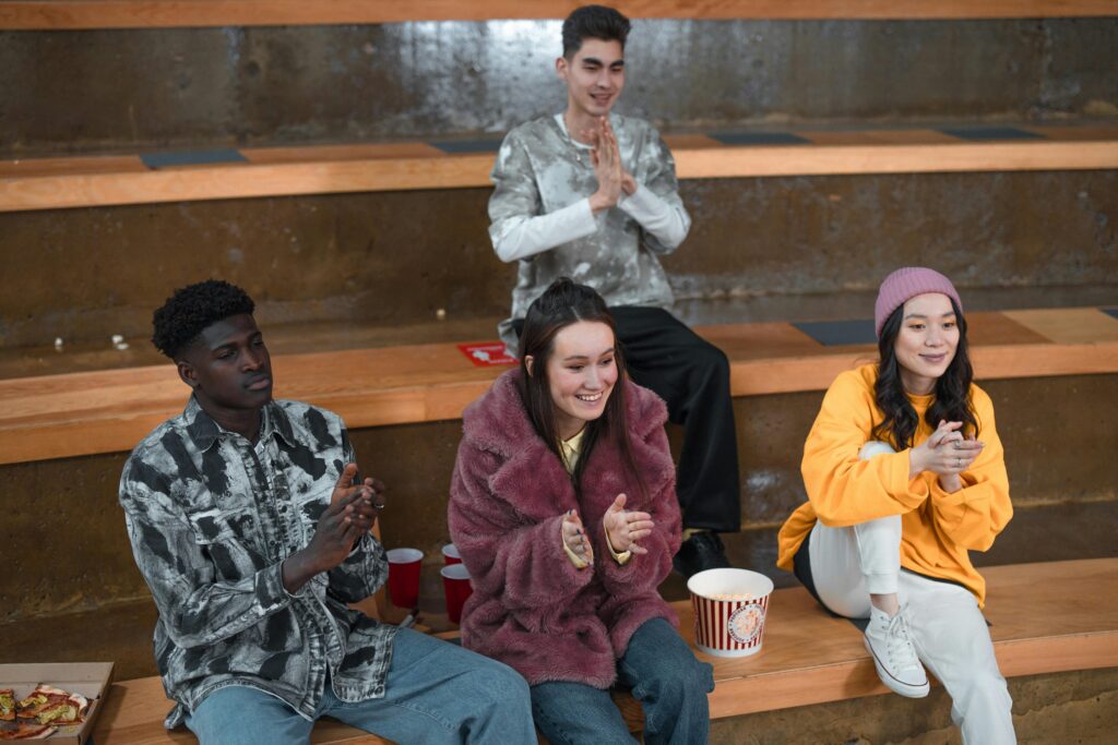 A diverse group of young adults sitting in bleachers, clapping and enjoying togetherness indoors.